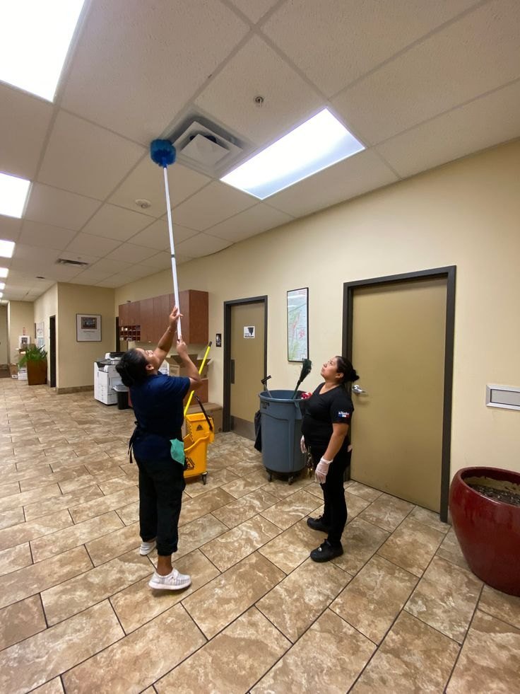 Two cleaners dusting the ceiling and vents in an office hallway with a long-handled duster
