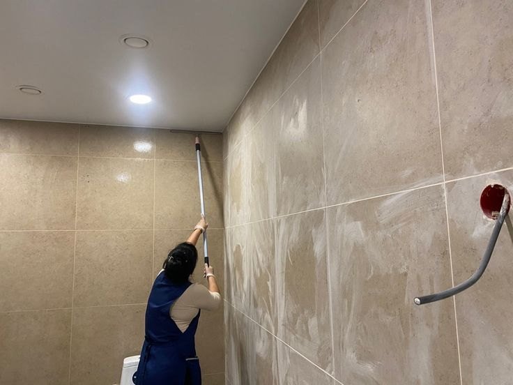 Cleaner using a long pole to wipe the tiled walls of an office restroom